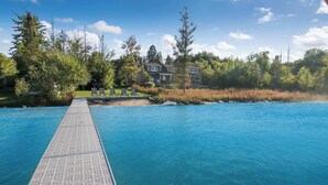 Eine hölzerne Mole führt zu einem klaren, blauen Pool, im Hintergrund ist ein Haus zu sehen.