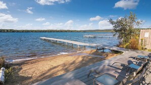 Eine Holzterrasse mit Liegestühlen mit Blick auf einen See, einen Sandstrand und einen Steg, der ins Wasser ragt.