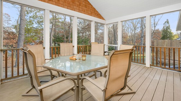 Eine überdachte Terrasse mit Glastisch und -stühlen, umgeben von großen Fenstern mit Blick auf Bäume und eine Holzterrasse.