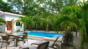 A swimming pool with lounge chairs and a table, surrounded by tropical plants and a white building with a red door. - Compass Rose House at Ceiba Beach Resort (Placencia)