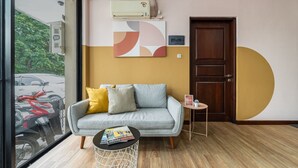 Ein modernes Hotelzimmer mit einer grauen Couch, einem kleinen Beistelltisch, einem großen Fenster mit Blick auf einen roten Roller und einer Wand-Klimaanlage.