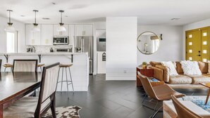A modern kitchen with white cabinets, a central island, and pendant lighting. There is a dining area with a wooden table and chairs, and a living room with a leather sofa and a round mirror. - Bello Grove | Old Town w/ Pool, Putting Green, Pinball & Games (Scottsdale)