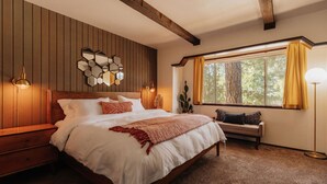 A bedroom with a wooden headboard, a large bed with white bedding, a bench with cushions, a wall-mounted hexagonal mirror, and a window with curtains. - Three bedroom  in United States of America, Big Bear (Big Bear)