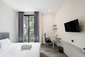 A modern hotel room with a large bed, a desk, a chair, a TV, and a view of greenery outside. - Cove Legacy (Jakarta)