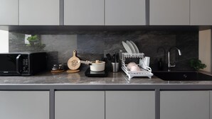 A modern kitchen with grey cabinets, a black microwave, a wooden cutting board, a white pot, a dish rack, and a sink. - Cove Legacy (Jakarta)