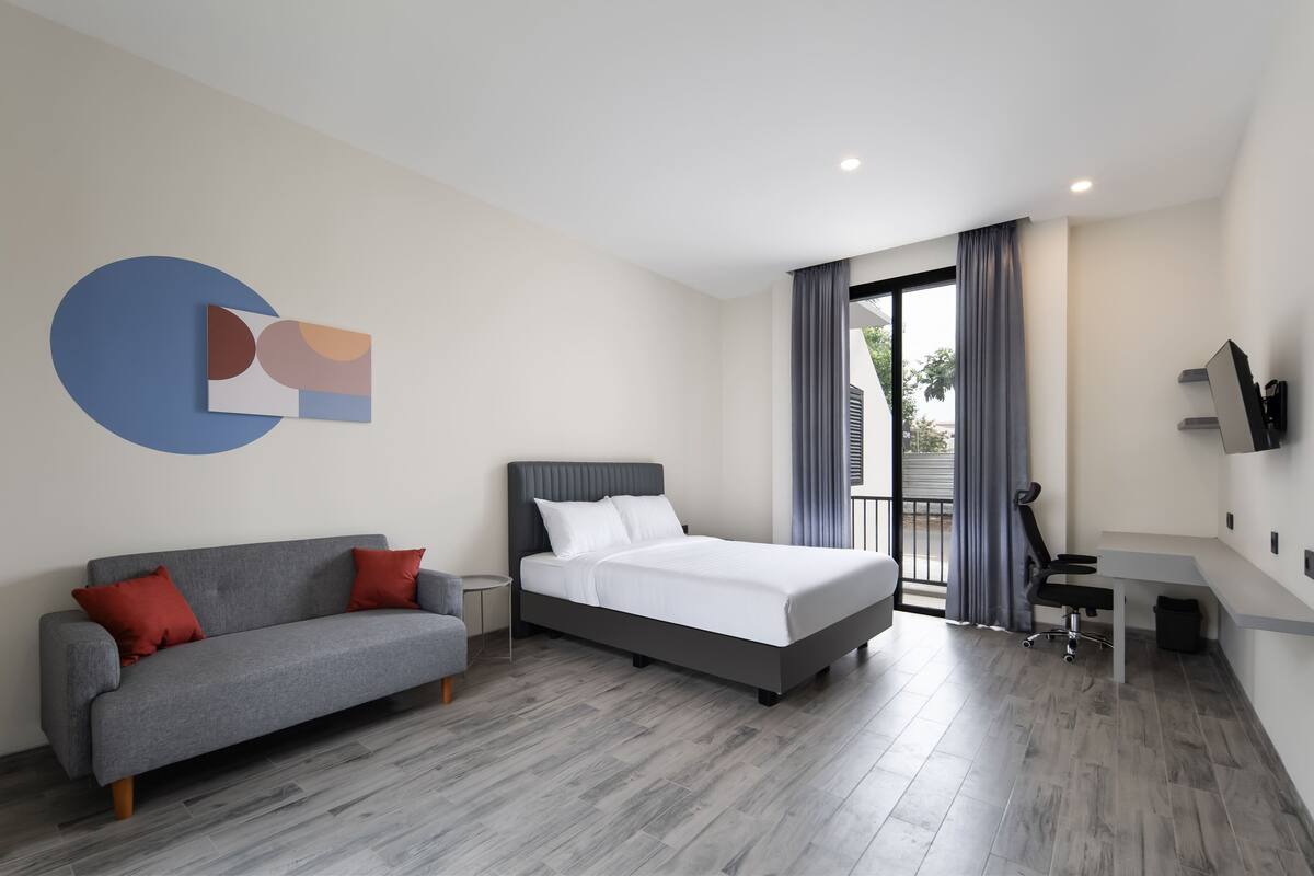 Quarto de hotel moderno com uma cama, um sofá cinzento, uma mesinha e uma televisão fixada na parede.