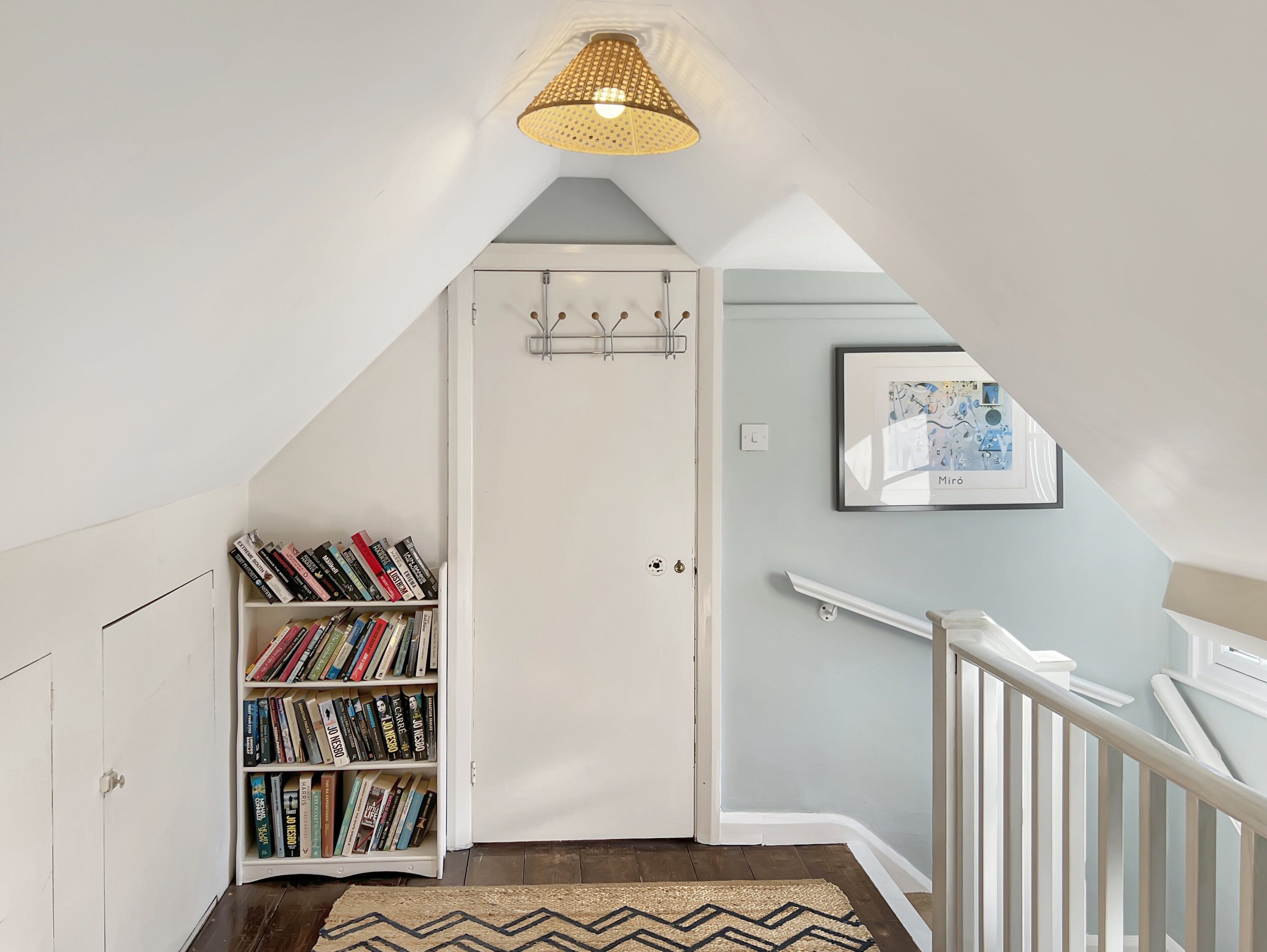 Un escalier avec une rampe blanche, une bibliothèque remplie de livres, une porte avec un porte-manteau et un luminaire suspendu.