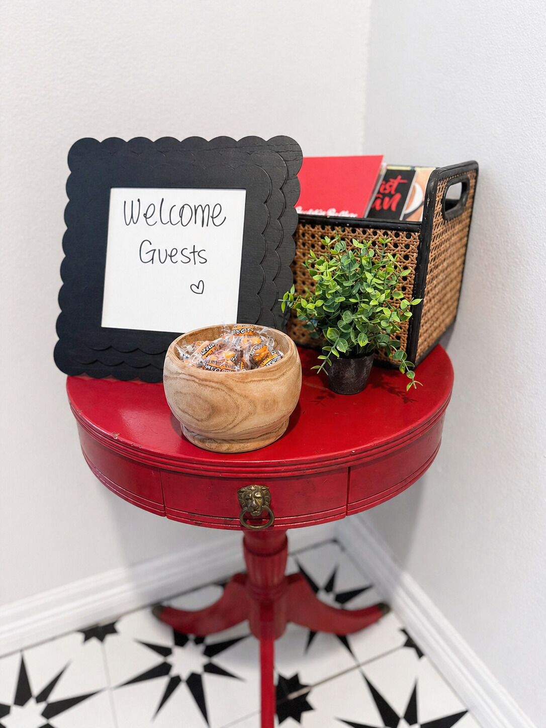 Une table rouge avec une pancarte de bienvenue, une petite plante et un panier rempli de collations.