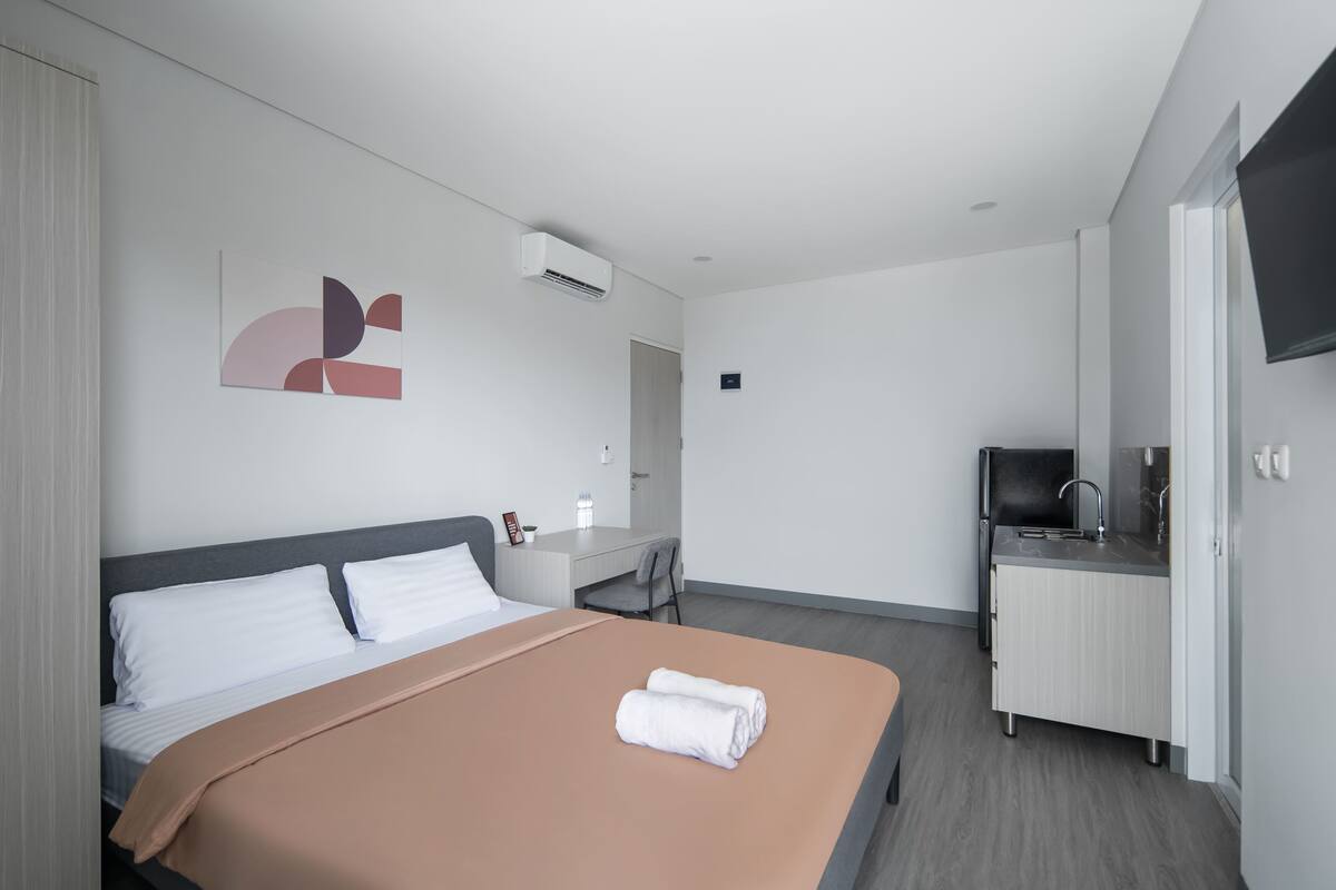 A modern hotel room with a bed, a desk, a kitchenette, and a flat-screen TV.