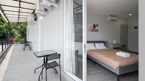 A modern hotel room with a bed, a small table, and chairs, and a balcony with a view of greenery. - Cove Anasera (South Tangerang)