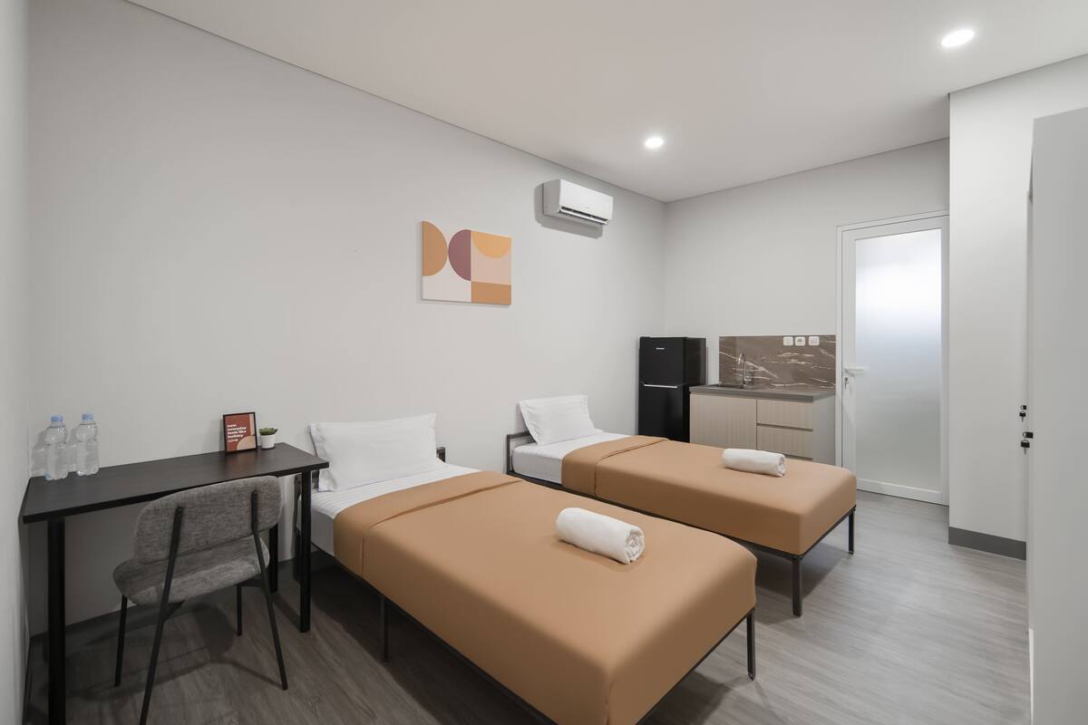 A hotel room with two beds, a desk, a chair, a small table with water bottles, a wall-mounted air conditioner, and a kitchen area with a refrigerator and sink.