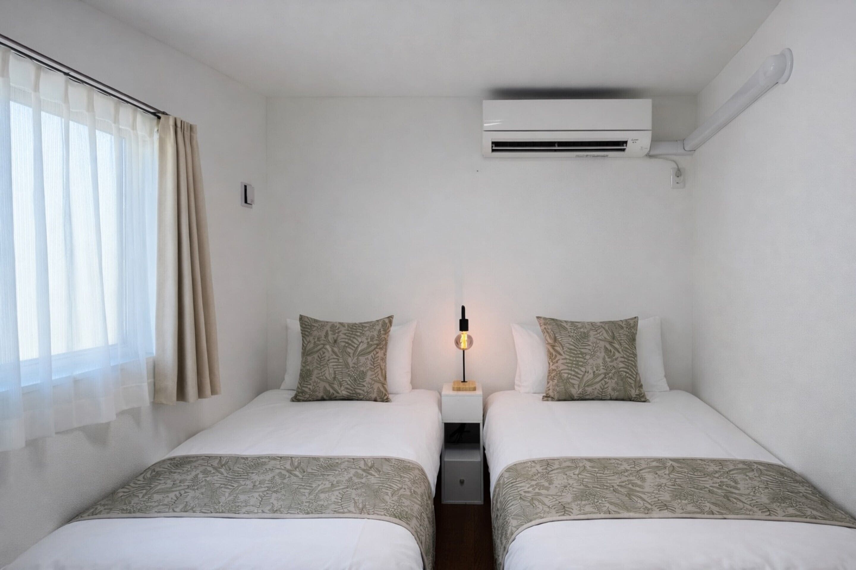 A hotel room with two beds, a nightstand, an air conditioning unit, and a window with curtains.