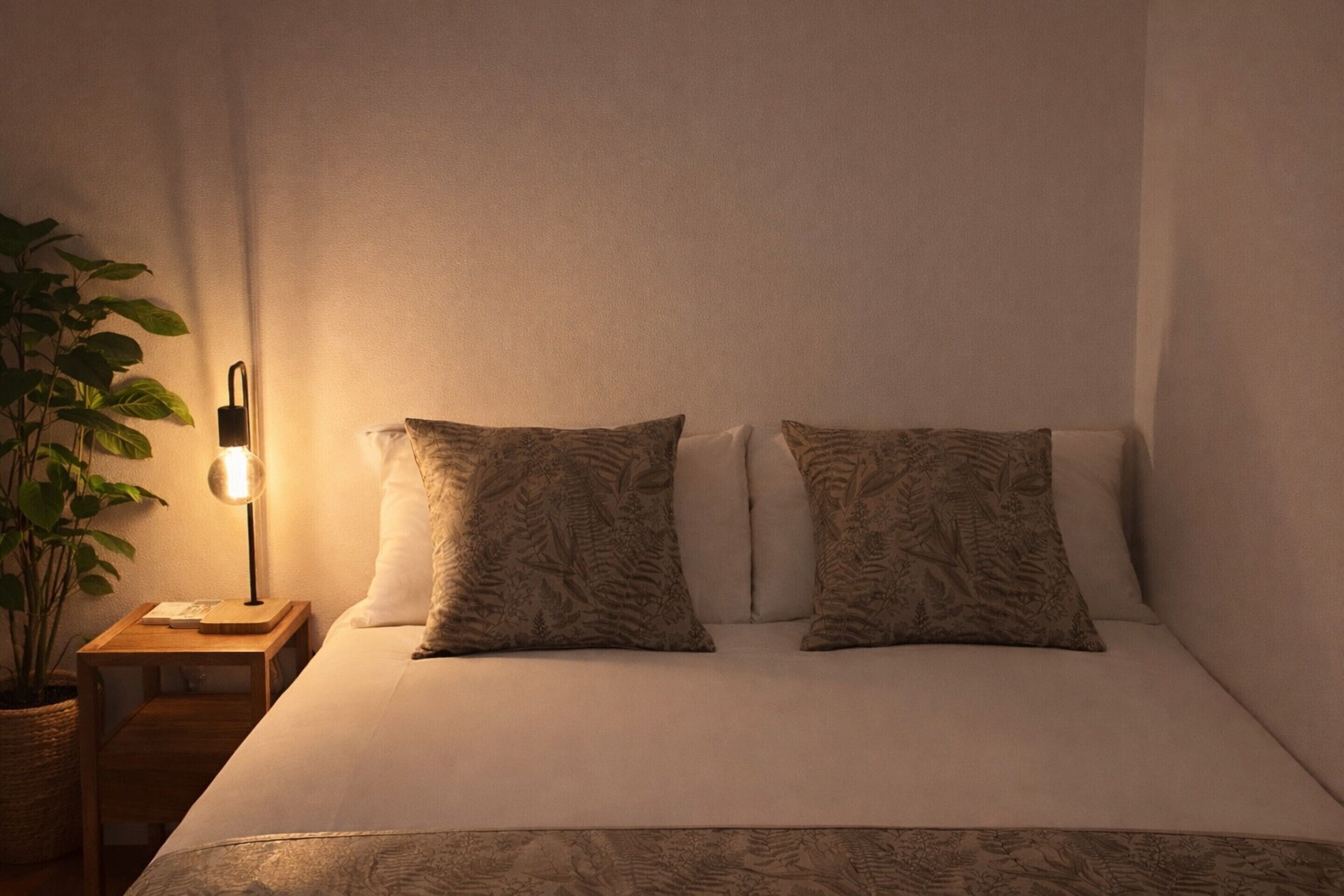 A neatly made bed with patterned pillows, a bedside lamp, and a potted plant on a wooden nightstand.