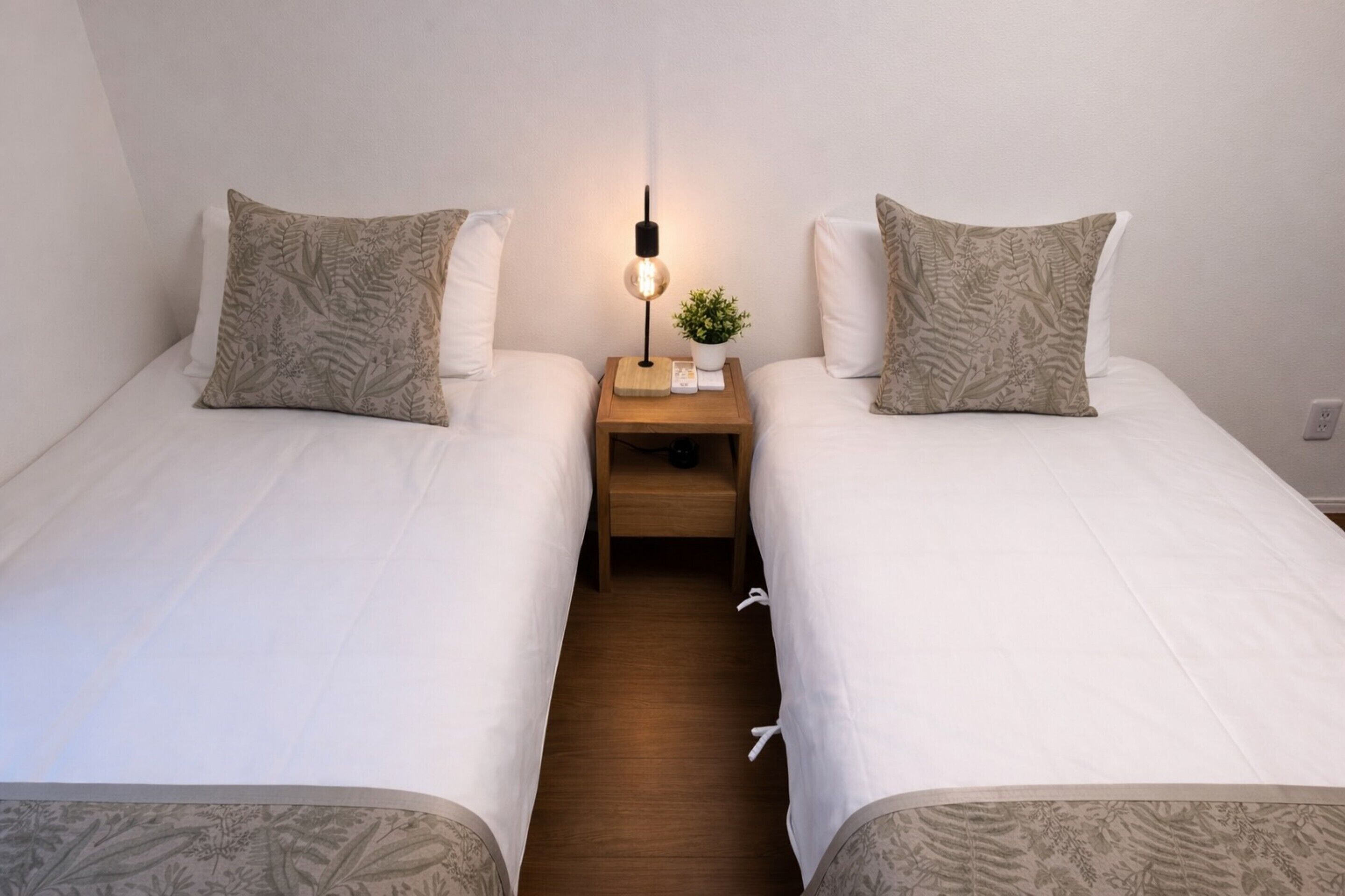 A twin room with two beds, a bedside table with a lamp, a small plant, and a wooden nightstand.