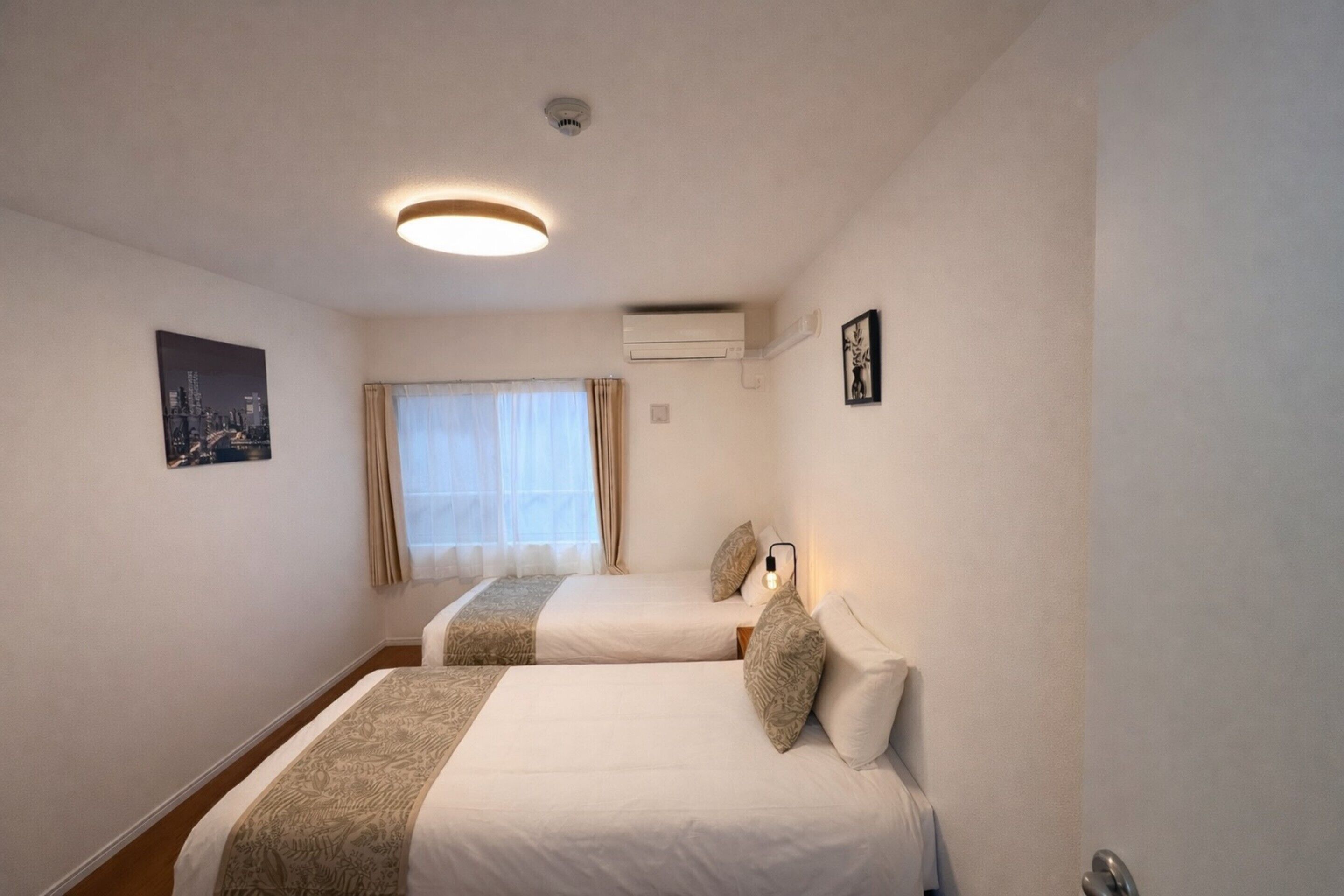 A hotel room with two beds, a window with curtains, an air conditioning unit, and a ceiling light.