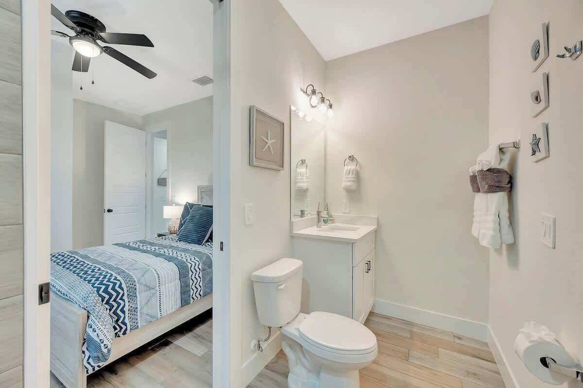 A bedroom with a bed, a ceiling fan, and a nightstand. The bathroom includes a toilet, sink, and towel rack.