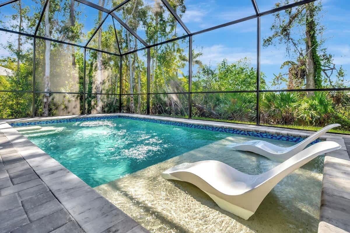 A glass-enclosed infinity pool with a white lounge chair, surrounded by lush greenery and trees.