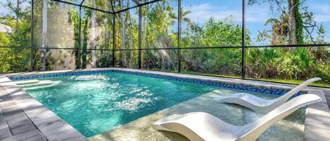 A glass-enclosed infinity pool with a white lounge chair, surrounded by lush greenery and trees.