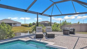 A covered outdoor swimming pool area with lounge chairs and a table, surrounded by a brick patio and a view of houses and greenery. - Coral Waters Modern Escape | Pool Home (Cape Coral)