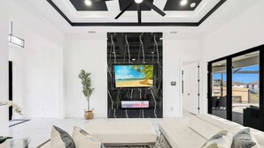 A modern living room with a large flat-screen TV mounted on a black marble wall, a seating area with a sofa and cushions, a potted plant, and a ceiling fan. - Coral Waters Modern Escape | Pool Home (Cape Coral)