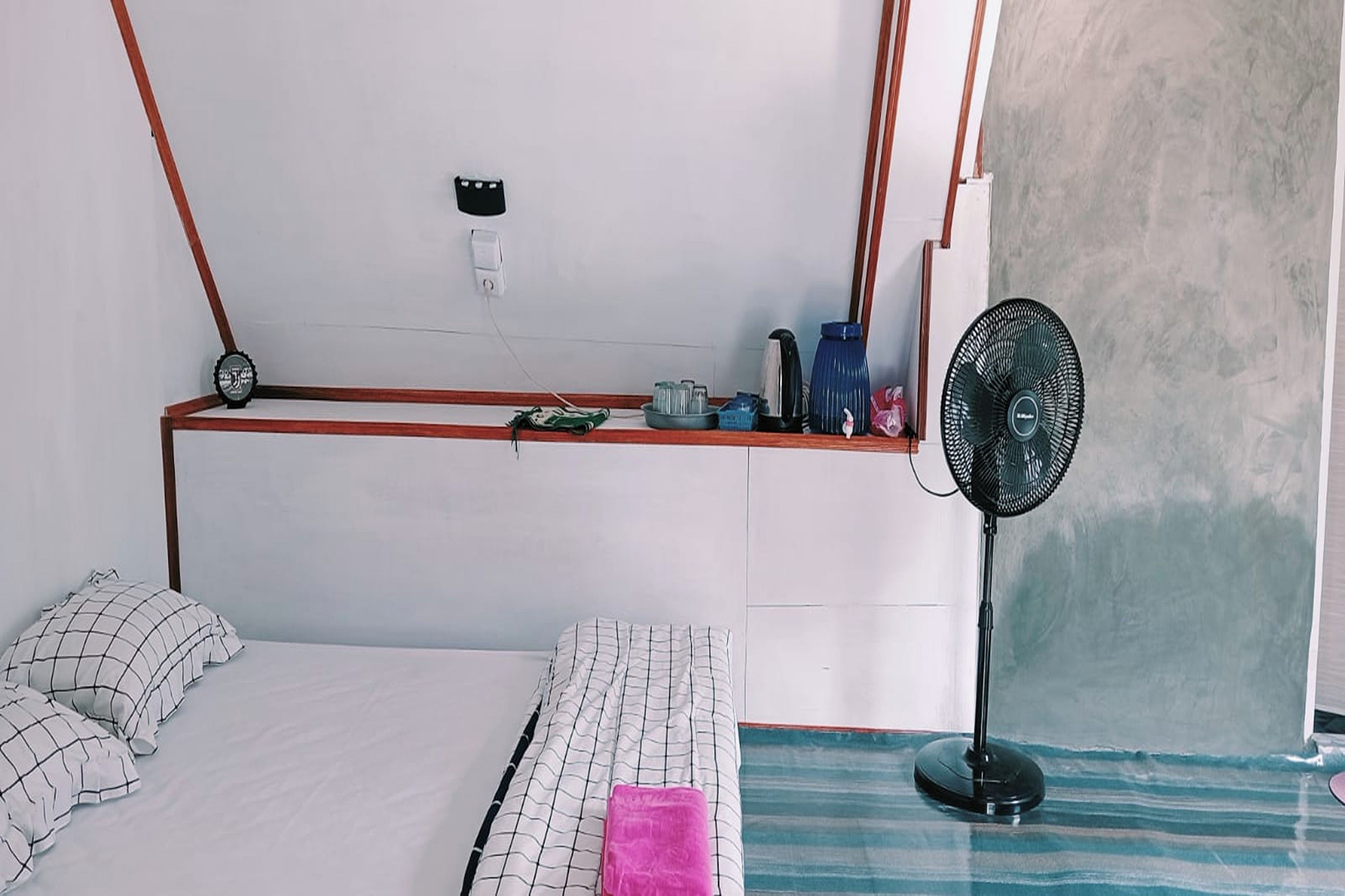 A bedroom with a bed, a bedside table, a fan, and a wall-mounted shelf with various items.