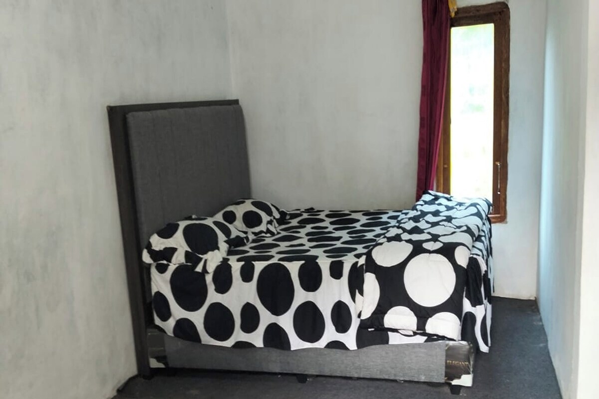 Een eenpersoonsbed met een zwart-wit stippen dekbed in een kamer met een raam en een deur.