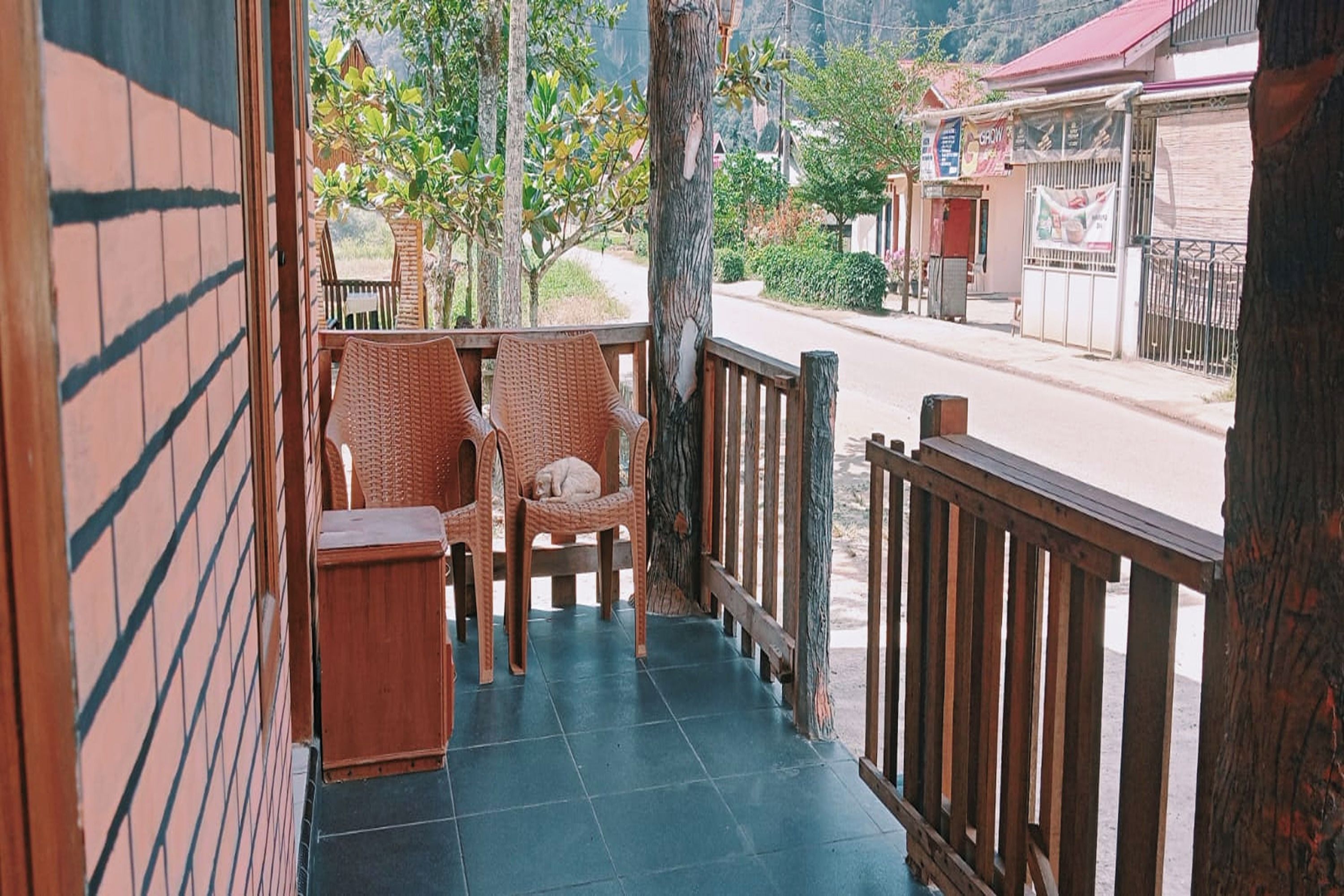 A balcony with wicker chairs and a small table, overlooking a street with buildings and trees.