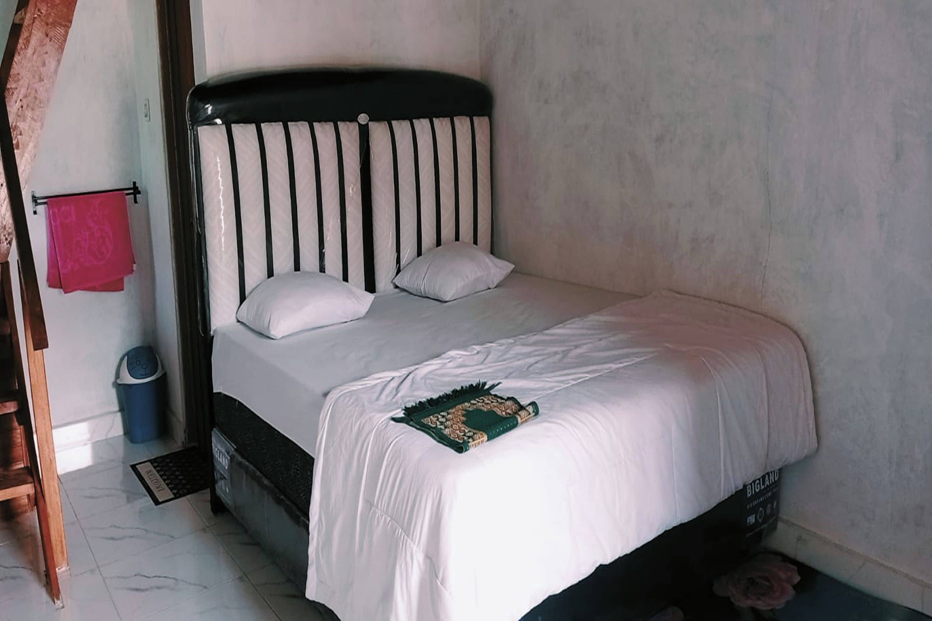 A bed with a black headboard and white bedding, a blue trash bin, and a red towel on a rack.