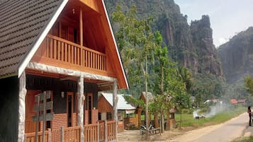 A wooden cabin with a balcony, surrounded by lush greenery and mountains.