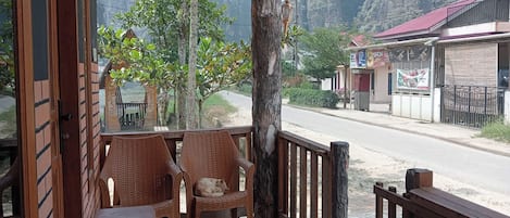 A wooden porch with two wicker chairs, a small table, and a view of a road and buildings in the background.
