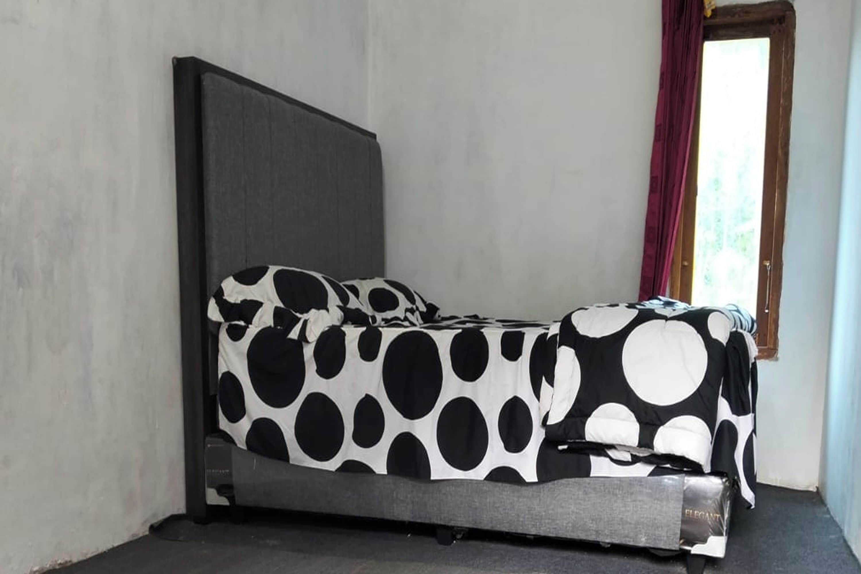 A bed with a black and white polka dot bedspread and a grey headboard in a room with plain walls and a window with red curtains.
