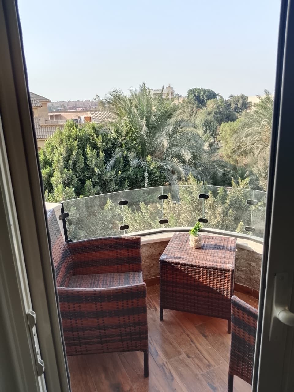 A balcony with wicker furniture and a potted plant, overlooking a lush green landscape with trees and buildings.