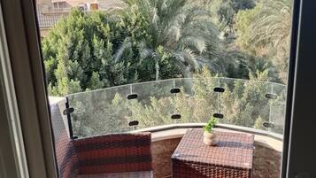 A balcony with wicker furniture and a potted plant, overlooking a lush green landscape with trees and buildings.