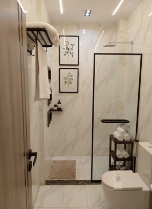 A modern bathroom with a glass shower enclosure, a toilet, and a small shelf with towels. - Zen Pyramids Luxury Villa (Giza)