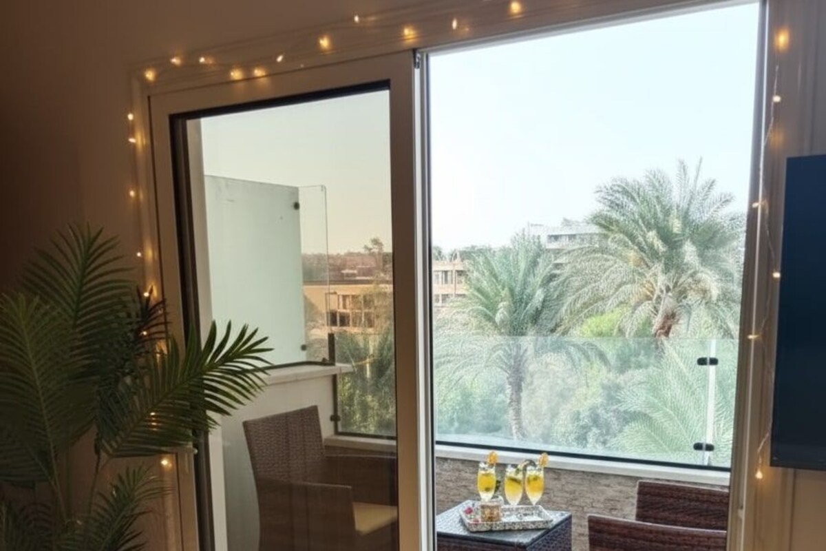 A room with a sliding glass door leading to a balcony with a view of palm trees and buildings. There is a wicker chair, a small table with drinks, and a potted plant.