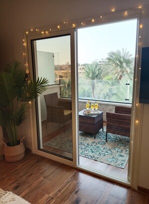 A room with a sliding glass door leading to a balcony with a view of palm trees and buildings. There is a wicker chair, a small table with drinks, and a potted plant. - Zen Pyramids Luxury Villa (Giza)