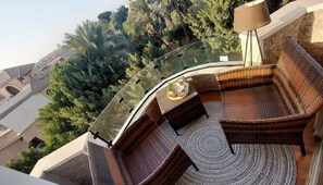 Un balcon doté de meubles en osier, d’une balustrade en verre et offrant une vue sur les bâtiments et les arbres environnants.