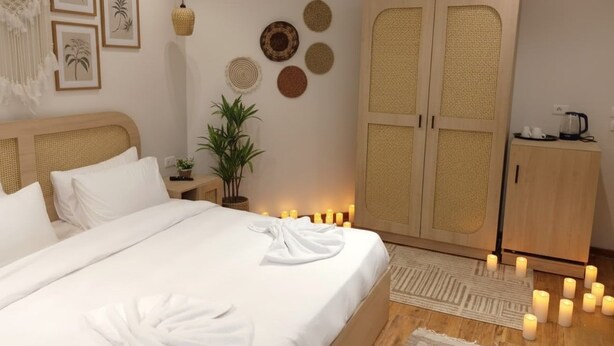 Ein ordentlich bezogenes Bett mit weißen Leinen und einem Handtuch, ein Holzschrank, ein kleiner Tisch mit einer Pflanze und Wanddekoration, darunter gerahmte Bilder und gewebte Körbe.