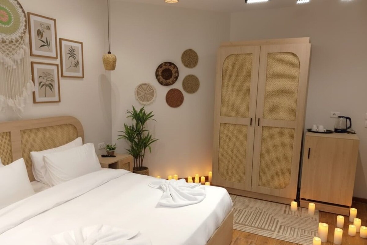 Ein ordentlich bezogenes Bett mit weißen Leinen und einem Handtuch, ein Holzschrank, ein kleiner Tisch mit einer Pflanze und Wanddekoration, darunter gerahmte Bilder und gewebte Körbe.