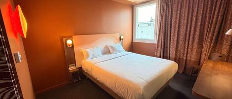 Habitación de hotel con una cama grande, una mesita de noche con lámpara, una ventana con cortinas y vistas a la ciudad.