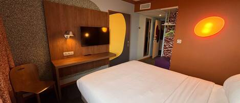 A hotel room with a bed, a desk with a TV, a mirror, and a shower area.