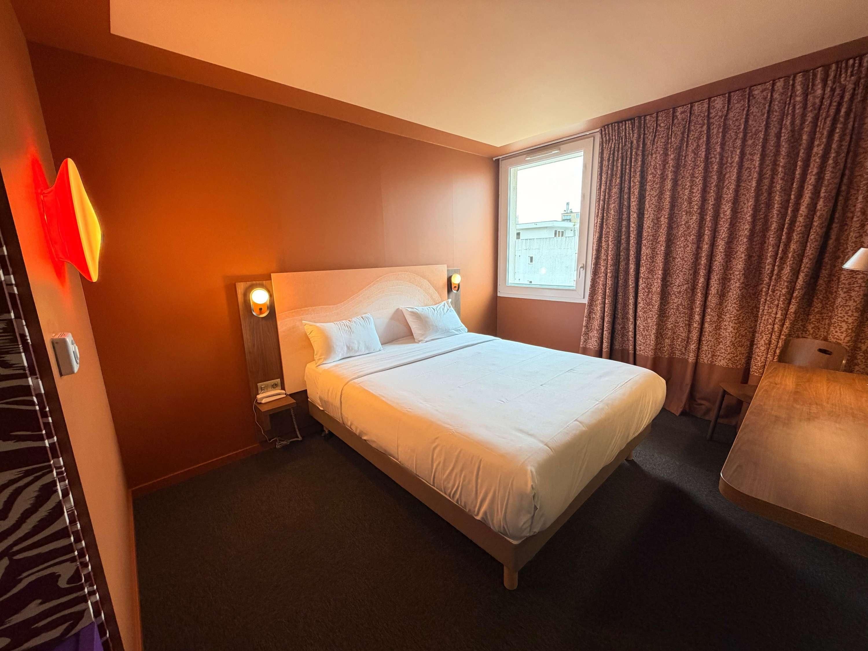 Habitación de hotel con una cama grande, una mesita de noche con lámpara, una ventana con cortinas y vistas a la ciudad.