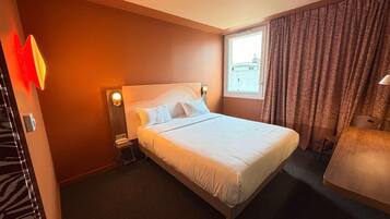 Habitación de hotel con una cama grande, una mesita de noche con lámpara, una ventana con cortinas y vistas a la ciudad.