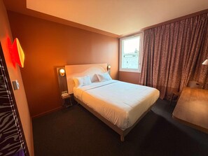 Habitación de hotel con una cama grande, una mesita de noche con lámpara, una ventana con cortinas y vistas a la ciudad.
