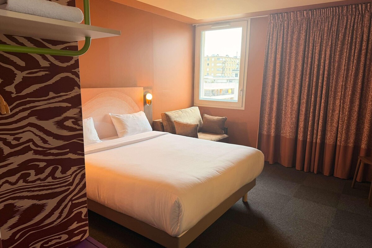 A hotel room with a large bed, a chair, a window with curtains, and a zebra-patterned wall.