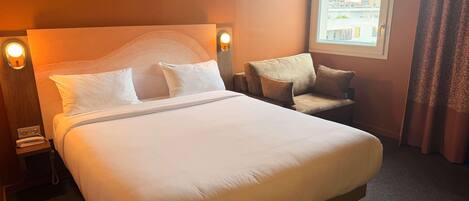 Habitación de hotel con una cama grande, un sofá, una mesita de noche con lámpara y una ventana con cortinas.