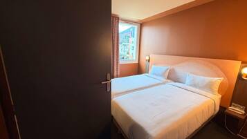 Quarto de hotel com uma cama, mesa de cabeceira, luminária e janela com cortinas.