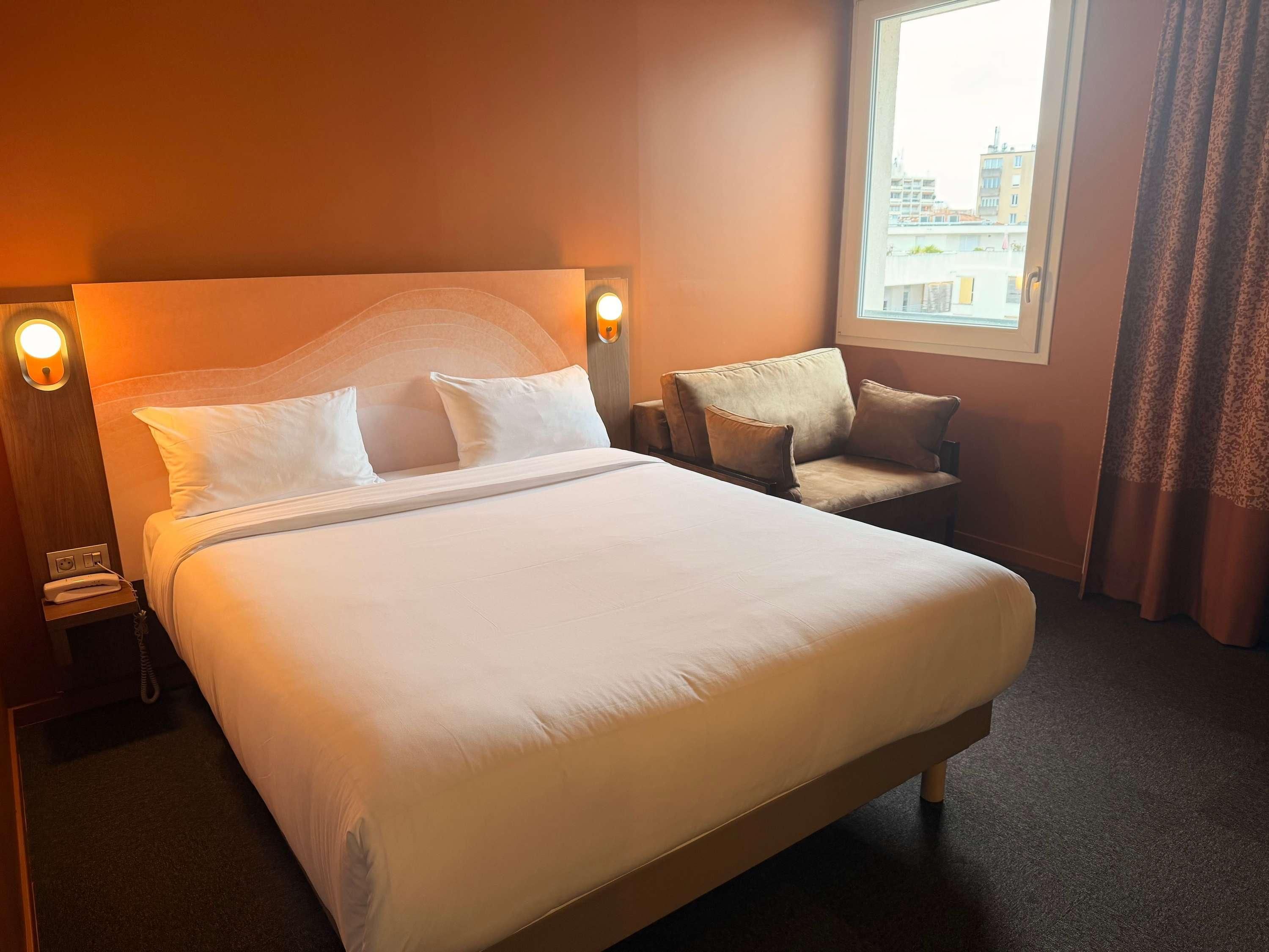 Ein Hotelzimmer mit einem großen Bett, einem Sofa, einem Nachttisch mit Lampe und einem Vorhangfenster.