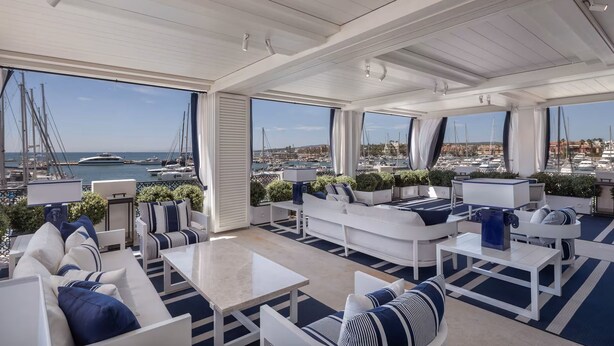 Un amplio espacio al aire libre con mobiliario blanco y detalles azules, con vistas a un puerto deportivo con barcos y un horizonte urbano.