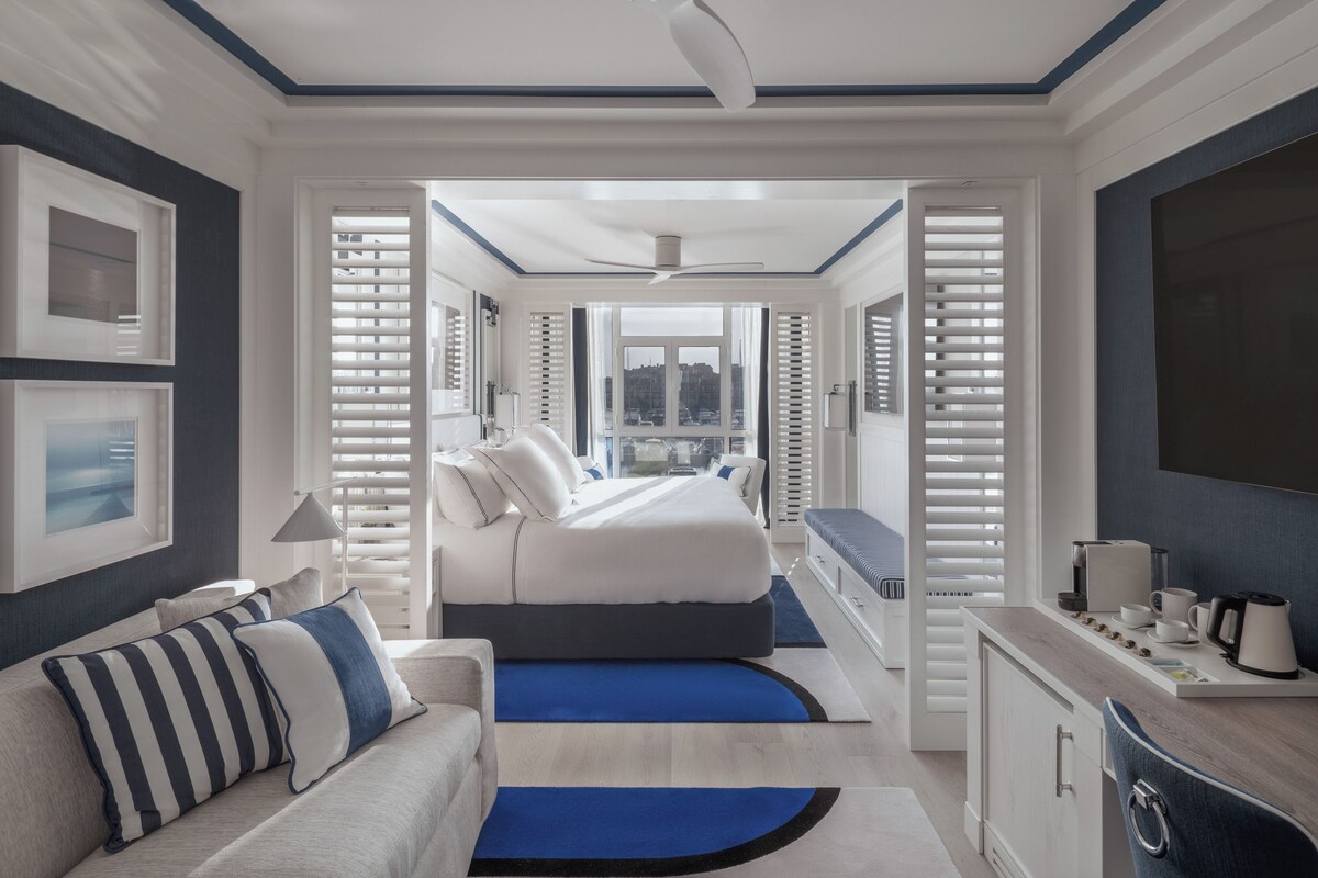 Une chambre d’hôtel moderne dotée d’un grand lit, d’un coin salon avec un canapé, d’un bureau avec une chaise et offrant une vue sur la ville par les fenêtres.